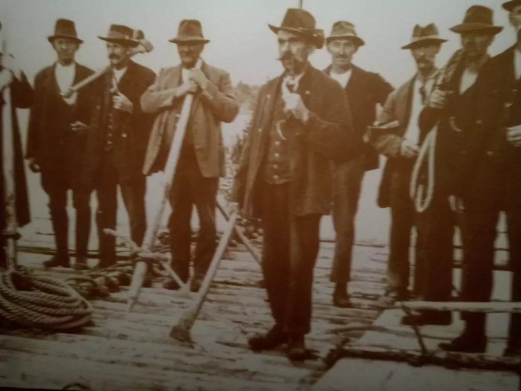 Eine Gruppe von Männern in altmodischer Kleidung und mit Hüten steht auf einem hölzernen Steg, einige halten Seile und Ruder.