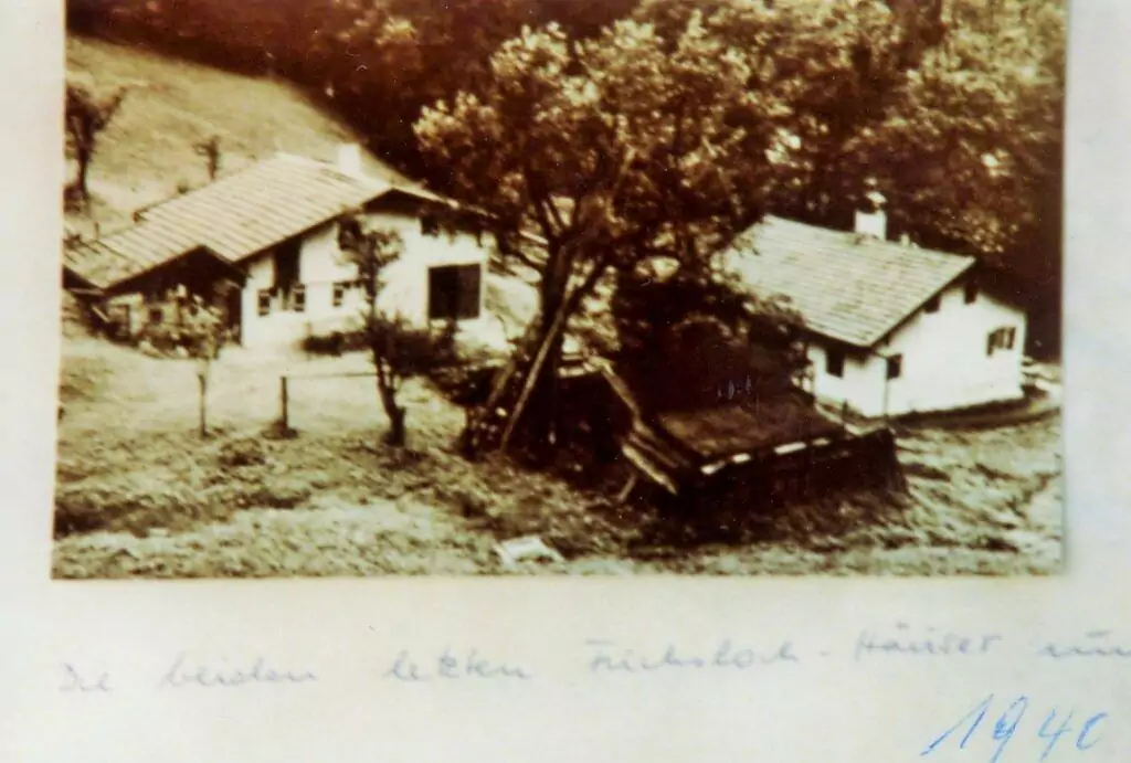 Eine sepiafarbene Fotografie von zwei kleinen Häusern, umgeben von Bäumen und Gras, mit handschriftlichem Text und der Jahreszahl 1946 am unteren Rand.