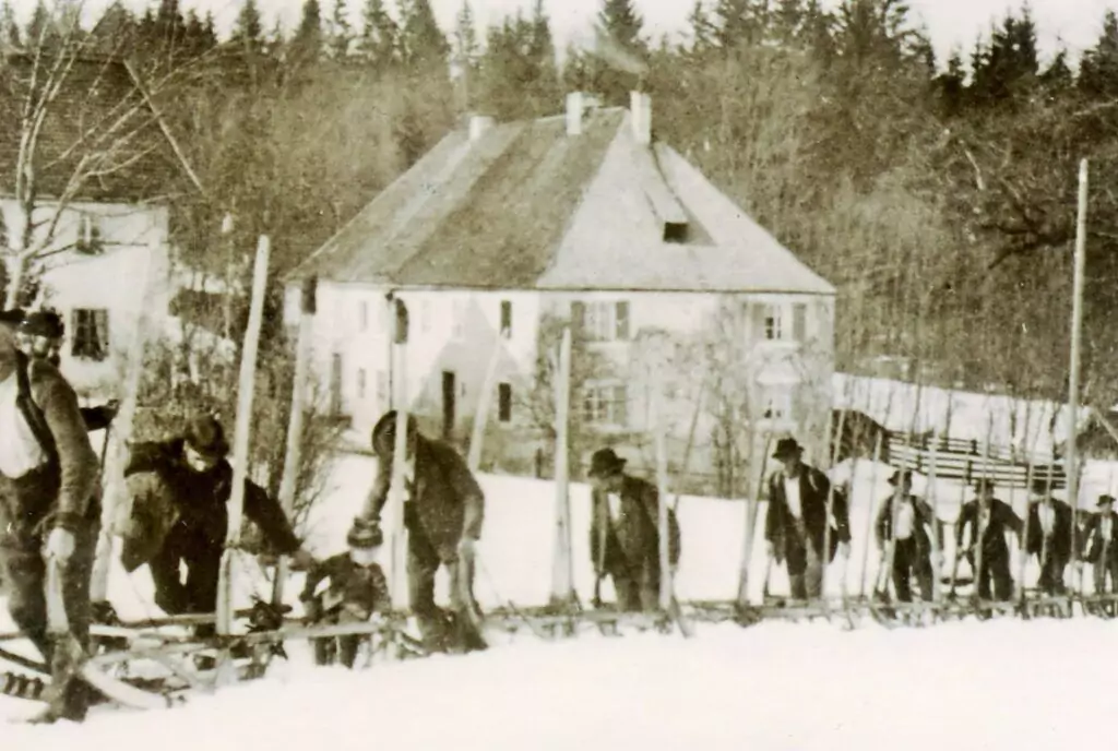 Eine Gruppe von Personen in Winterkleidung geht mit Skiern und einem Hornschlitten auf Schnee bergauf, vorbei an einem großen, von Bäumen umgebenen Haus.