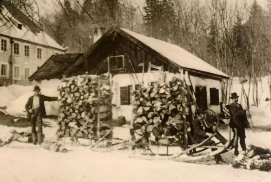 Zwei Männer stehen neben Hornschlitten, die mit gehacktem Brennholz beladen sind, vor einer Holzhütte in einer verschneiten, ländlichen Landschaft mit Bäumen und Gebäuden im Hintergrund.