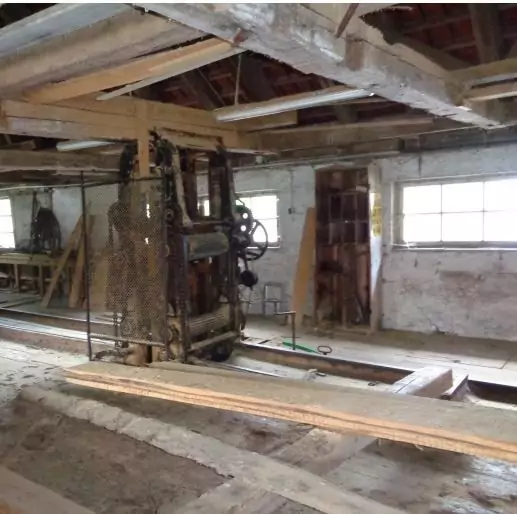 Das Innere eines alten Sägewerks mit Holzbalken, einer großen mechanischen Säge und natürlichem Licht, das durch die Fenster auf der rechten Seite einfällt.