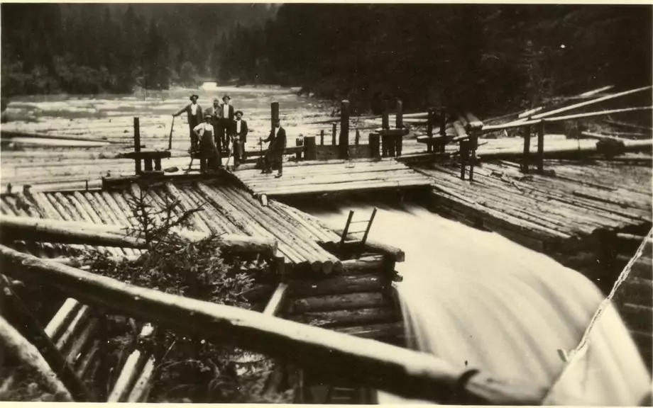 Eine Gruppe von Menschen steht auf einer Holzkonstruktion über dem fließenden Fluss Trift, umgeben von Baumstämmen und einer bewaldeten Landschaft im Hintergrund.