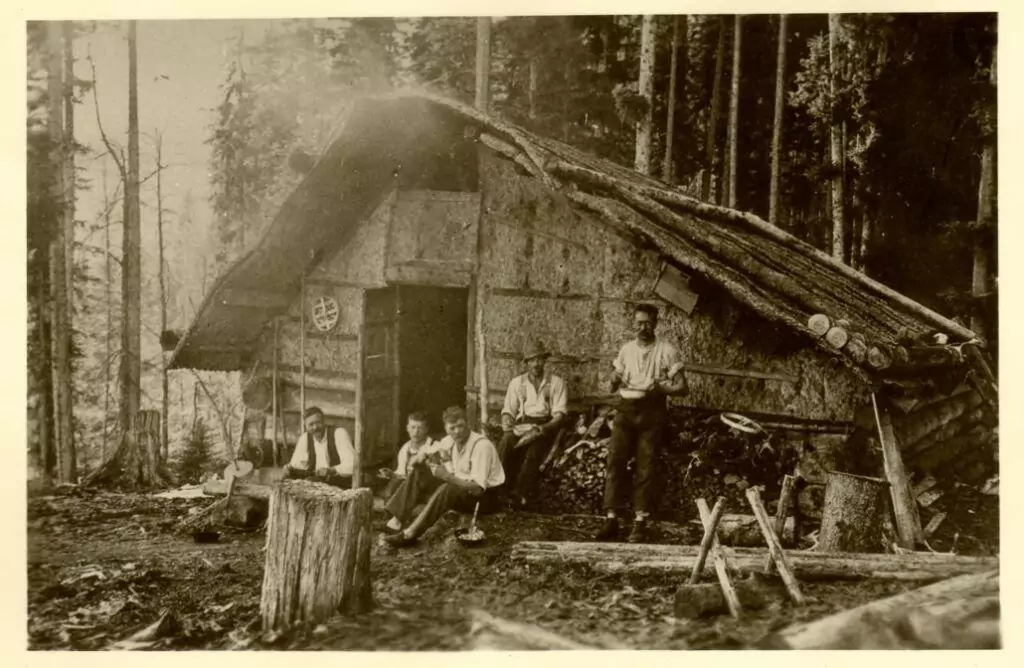 Fünf Personen sitzen und stehen vor einer rustikalen Hütte in einem Wald, in dem Werkzeuge und Holzscheite im Vordergrund verstreut sind.