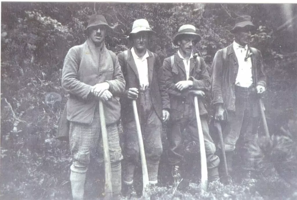 Vier Männer in Outdoor-Kleidung und Hüten stehen nebeneinander in der Natur und halten jeweils ein langstieliges Werkzeug, möglicherweise für die Land- oder Forstwirtschaft.