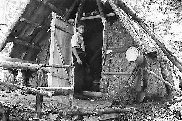 Ein Junge steht am Eingang einer rustikalen Holzhütte mit Strohdach, umgeben von Bäumen und grob behauenen Stämmen.