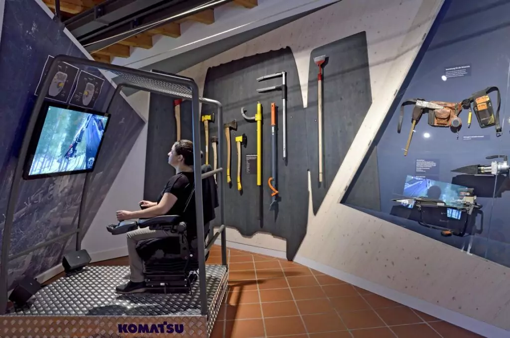 Eine Person bedient einen Komatsu-Simulator in einer Ausstellung des Holzknechtmuseums Ruhpolding, in der forstwirtschaftliche Werkzeuge und Maschinen an der Wand gezeigt werden.