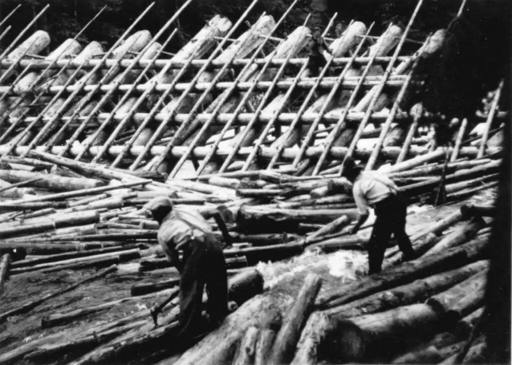 Zwei Arbeiter stehen zwischen schwimmenden Baumstämmen an der Trift, während die Stämme diagonal auf einer Holzkonstruktion im Hintergrund angeordnet sind.