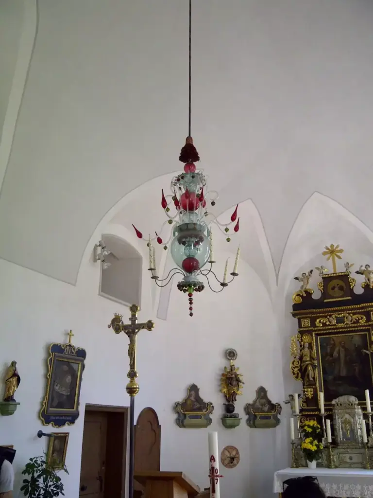 Ein gläserner Kronleuchter mit roten Akzenten hängt von der Decke eines Kirchenraums mit religiösen Gemälden, Statuen, einem Kruzifix und einem verzierten Altar.