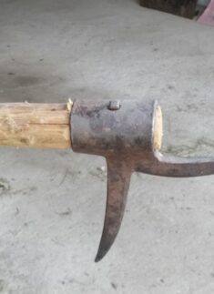 Nahaufnahme eines abgenutzten, zweizinkigen Holzerwerkzeug-Metallspitzkopfes, der an einem rauen Holzstiel befestigt ist und auf einer Betonfläche ruht.