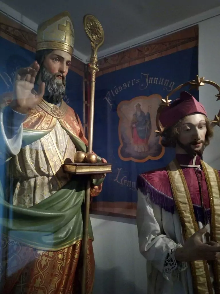 Zwei religiöse Statuen in prächtigen Gewändern, von denen eine einen Stab und die andere eine Sternenkrone trägt, sind vor einem blau-goldenen Banner aufgestellt.