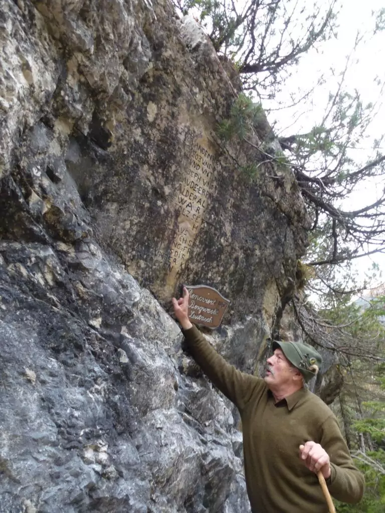 Eine Person in Outdoor-Kleidung zeigt auf eine eingravierte Inschrift auf einer Felswand und hält ein Holzschild mit der Aufschrift "Loiten und Riesen Inscript Steingravur engraved rockwall".