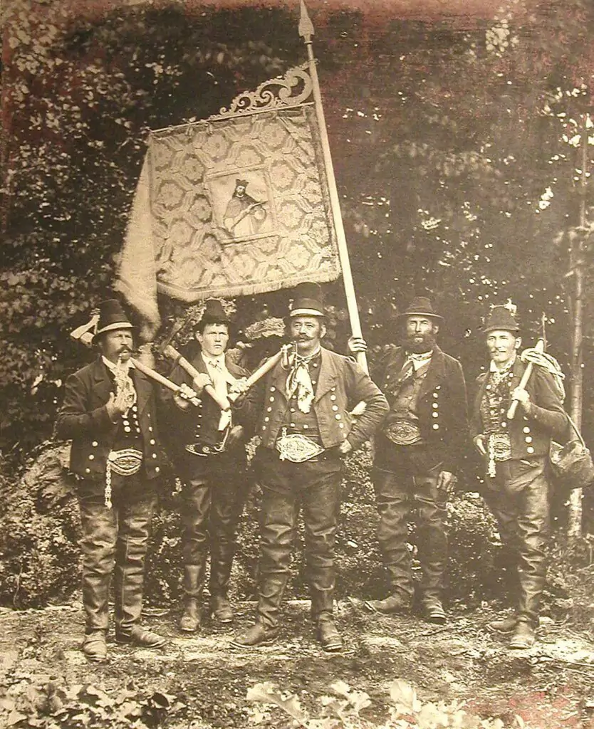 Fünf Männer in traditioneller Kleidung und mit Hüten stehen im Freien, halten Gewehre und eine geschmückte Fahne in der Hand und posieren für ein altes Schwarz-Weiß-Foto.