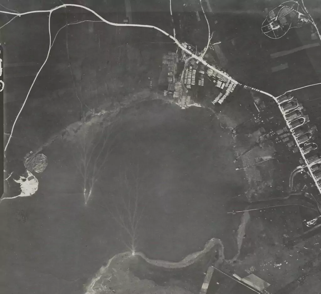 Schwarz-Weiß-Luftaufnahme des Küstengebiets von Trift mit Wasser, Uferlinie, Docks, Straßen, verstreuten Gebäuden und Booten mit sichtbaren Wellen im Wasser.