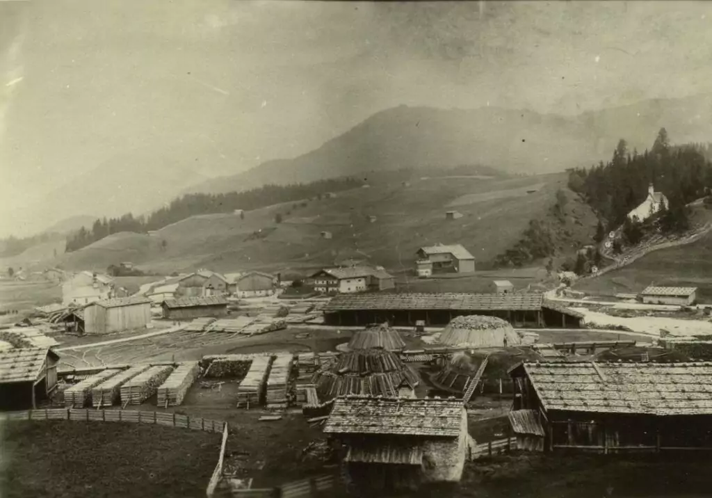 Schwarz-Weiß-Foto eines ländlichen Dorfes mit Holzhäusern und Zäunen in einer hügeligen Landschaft mit Bergen im Hintergrund.