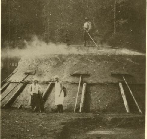 Drei Personen arbeiten um einen großen, rauchenden Holzkohleofen aus Erde und Holz, der sich im Freien in einem Waldgebiet befindet.