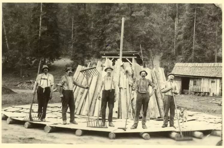 Fünf Männer stehen auf einem Holzfloß und halten Werkzeuge in der Hand, während im Hintergrund eine Holzkonstruktion und ein Wald zu sehen sind.