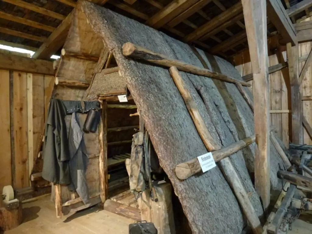 Ein rustikaler hölzerner Kobel mit schrägem Dach, der aus Baumstämmen und Rinde gebaut wurde, steht im Innenraum, wo Kleidung und Ausrüstung an der Seite aufgehängt sind, was an die traditionelle Hüttenarbeit erinnert.