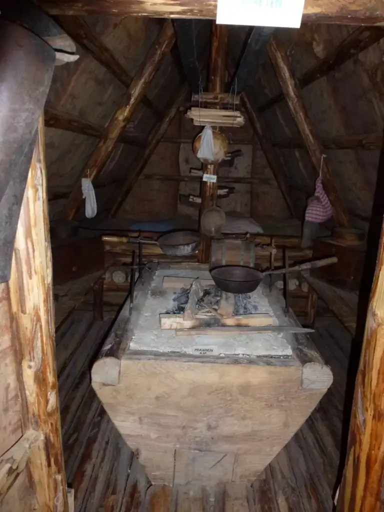 Das Bild zeigt das Innere einer rustikalen Holzhütte mit einem zentralen Steinofen, Töpfen, Holzmöbeln und hängenden Utensilien.