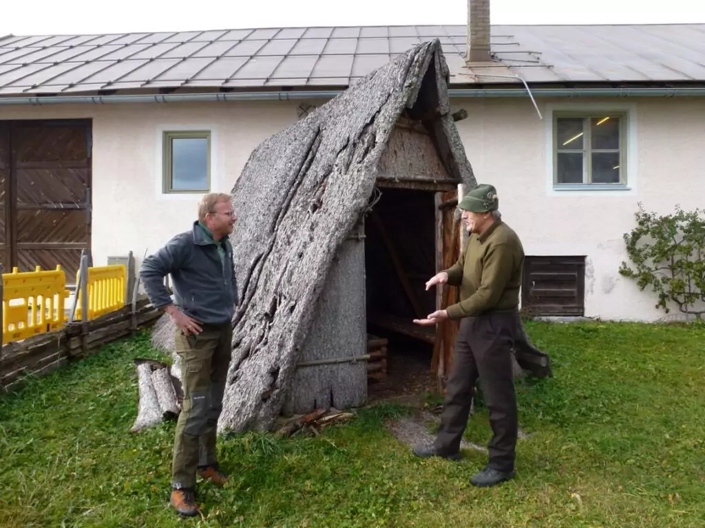 Zwei Männer stehen vor einer kleinen Holzhütte mit schrägem Rindendach und unterhalten sich in der Nähe des Kulturhauses Leutasch, vor einem größeren Gebäude.