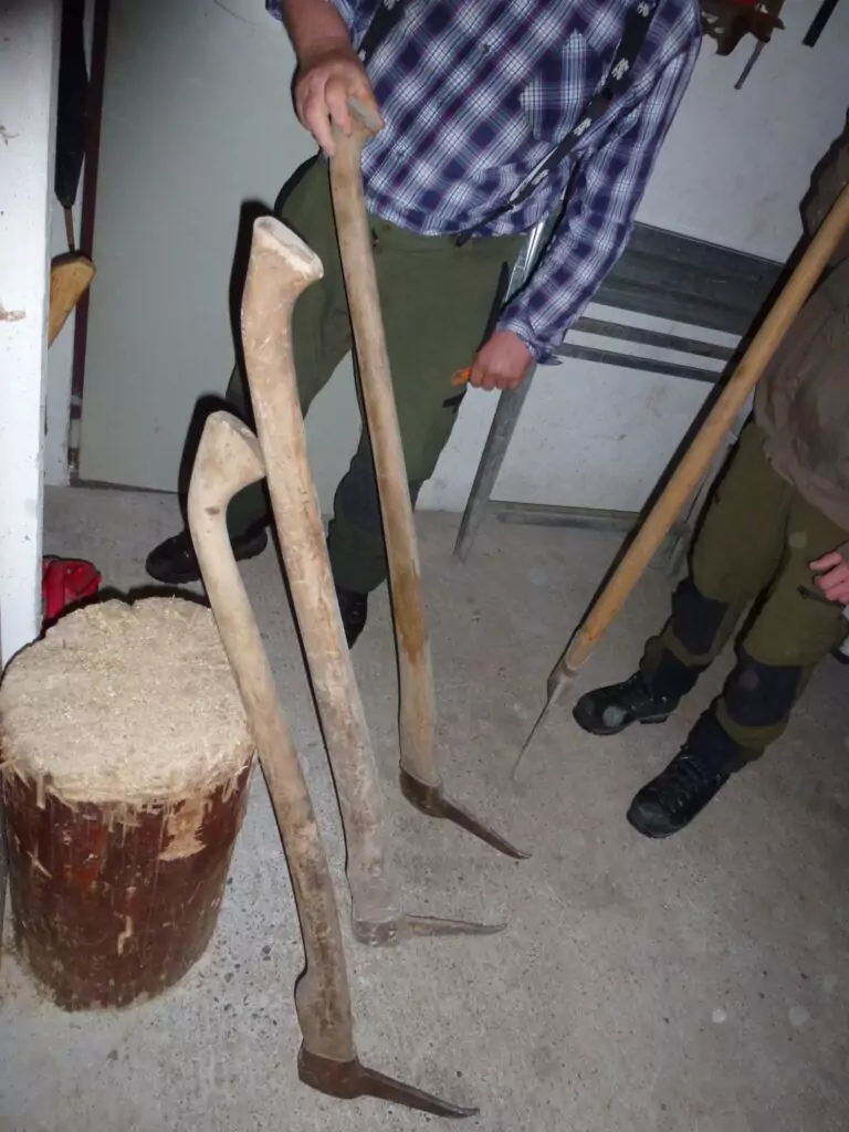 Zwei Personen stehen drinnen neben drei Holzerwerkzeugen mit langen gebogenen Stielen und spitzen Metallspitzen, die an einen Hackblock auf dem Betonboden gelehnt sind.