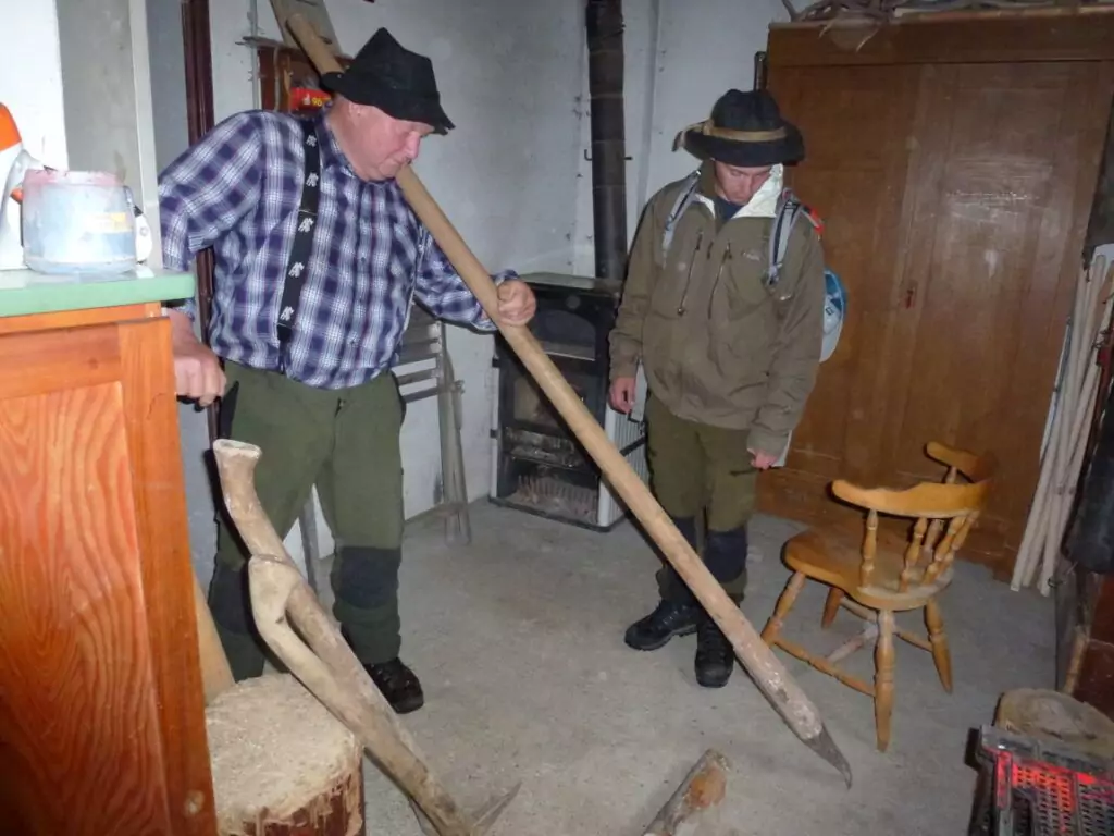Zwei Männer in traditioneller Kleidung stehen in einem Haus; einer bläst in ein langes hölzernes Alphorn, während der andere zuschaut. Im Hintergrund sind Holzerwerkzeuge und Holzmöbel sowie ein Ofen zu sehen.