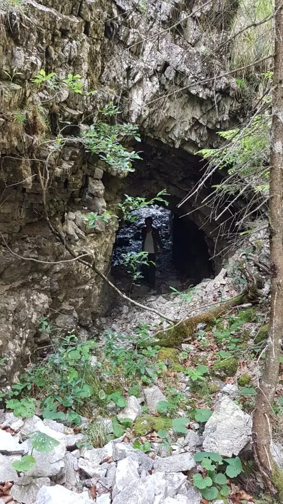 Felsiger Höhleneingang, umgeben von Vegetation und Bäumen, mit verstreuten Steinen und Grünzeug auf dem Boden - angeblich ein versteckter Treffpunkt für Loiten und Riesen.