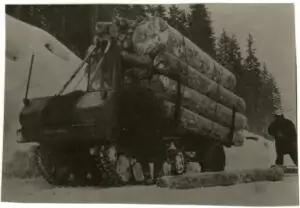 Ein mit großen Baumstämmen beladenes Raupenfahrzeug ist auf einer verschneiten Straße in der Nähe eines Waldes abgestellt, während eine Person in der Nähe steht und einen Baumstamm bearbeitet.