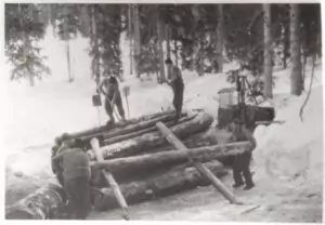 Vier Personen manövrieren mit Werkzeugen große Stämme in einem verschneiten, von Bäumen umgebenen Wald, während Raupenfahrzeuge bereitstehen, um beim Heben schwerer Lasten zu helfen.