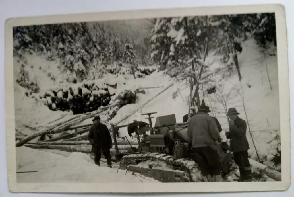 Eine Gruppe von Männern steht neben einem Traktor und gefällten Baumstämmen in einem verschneiten Wald. Im Hintergrund sind Raupenfahrzeuge und gestapelte Baumstämme zwischen den Bäumen zu sehen.