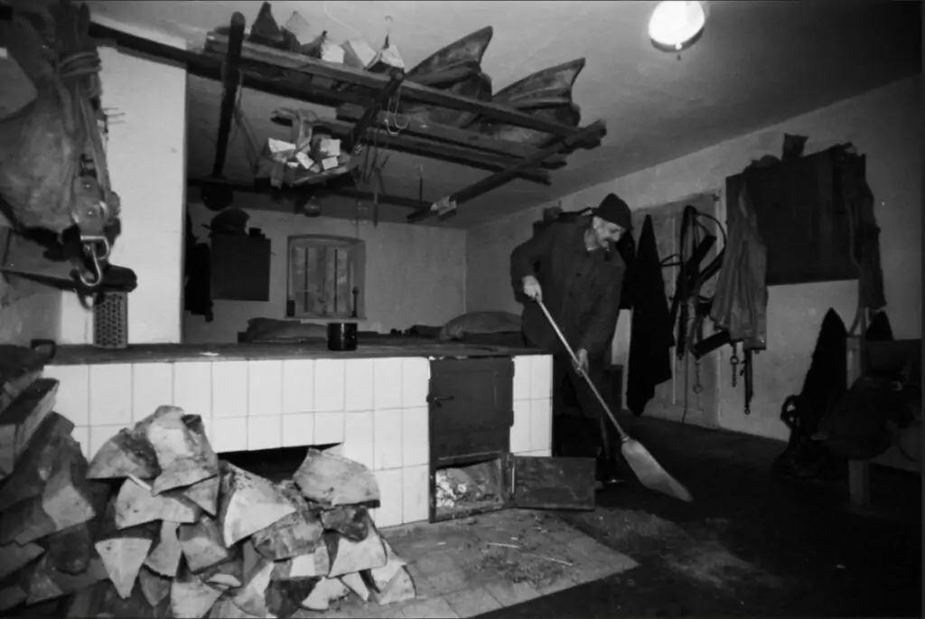 Eine Person in Mantel und Hut schaufelt neben einem Kachelofen in einem Raum im Hüttenstil, der mit Brennholz, Kobel-Werkzeugen und an den Wänden und der Decke hängenden Geräten vollgestopft ist.