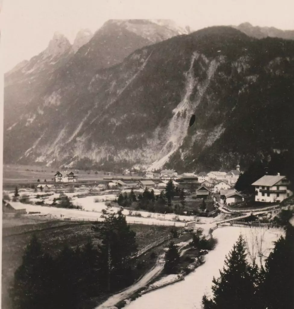 Schwarz-Weiß-Foto eines Dorfes mit verstreuten Häusern, einem Fluss und Bergen im Hintergrund.
