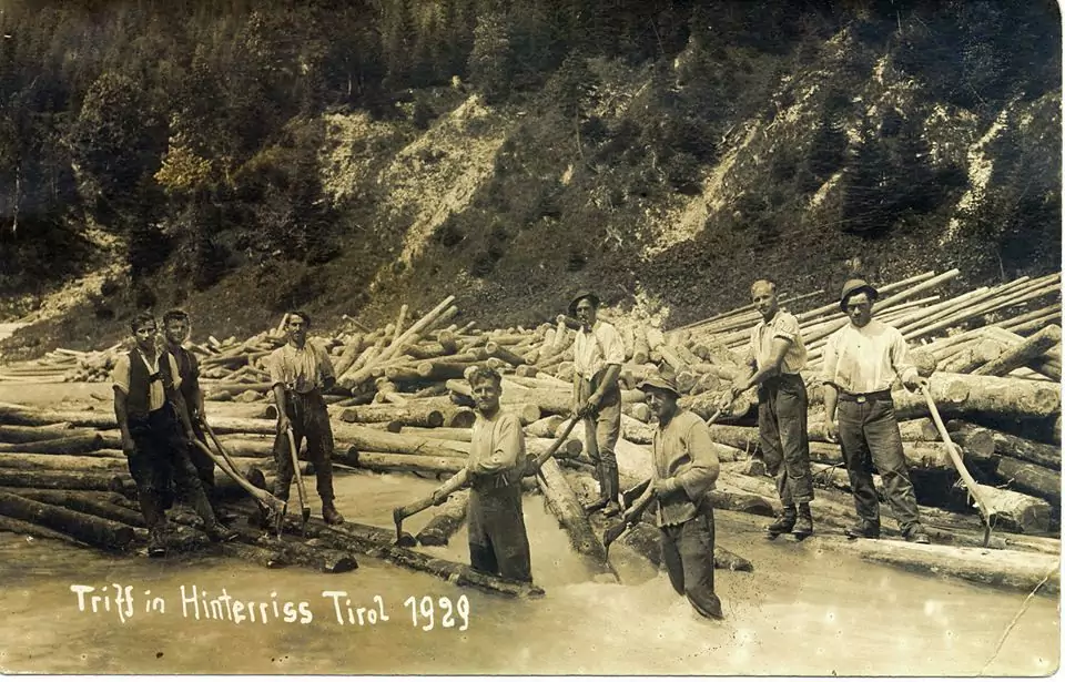 Schwarz-Weiß-Foto von neun Männern, die auf schwimmenden Baumstämmen während einer Trift in einem Fluss stehen, mit bewaldeten Hügeln im Hintergrund. Der Text lautet: "Trift in Hinterriss Tirol 1929.
