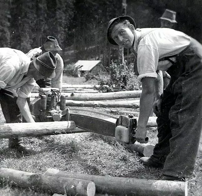 Zwei Männer sägen mit einer großen mechanischen Holzerwerkzeugsäge Holzstämme im Freien, während im Hintergrund weitere Stämme und Menschen zu sehen sind.