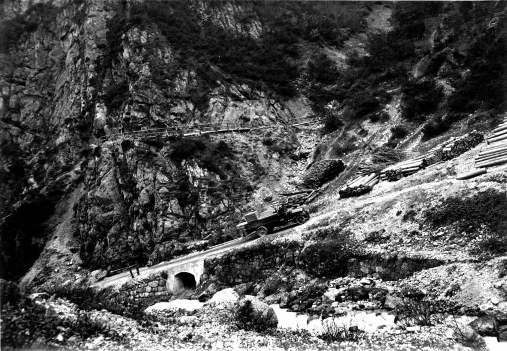 Ein Lastwagen fährt über eine schmale Bergstraße mit einer Steinbrücke, umgeben von steilem, felsigem Gelände und verstreuten Baumstämmen.