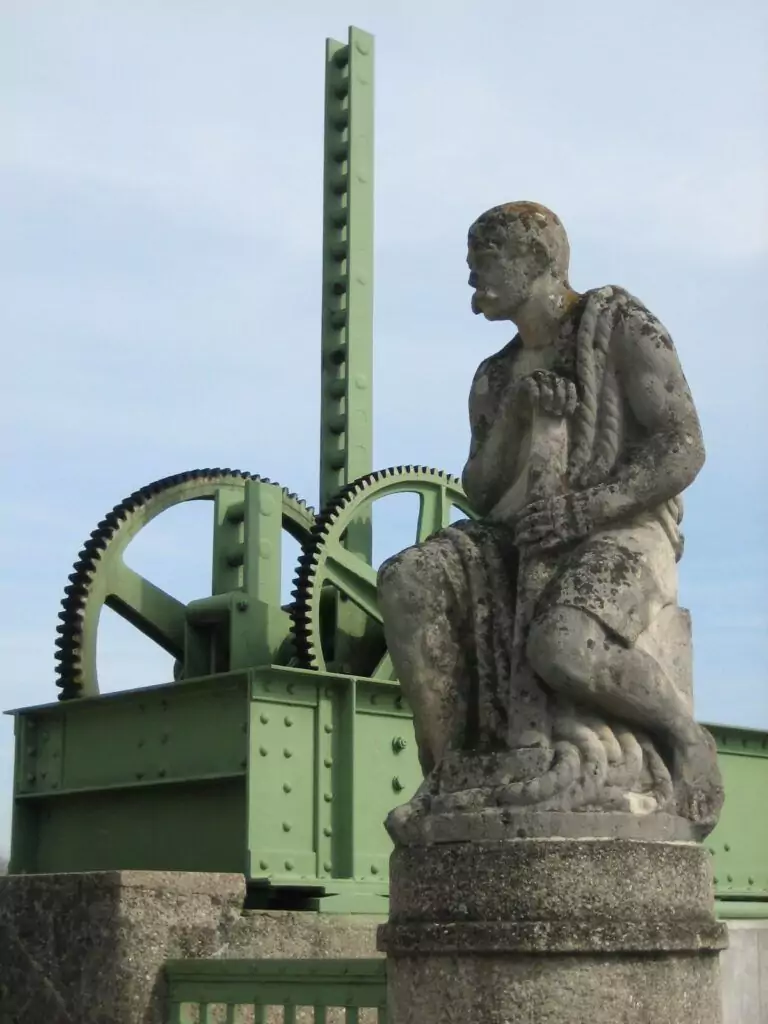 Eine verwitterte Steinstatue eines sitzenden Mannes, der ein Seil hält, ist eines der Denkmäler der Region und steht vor einer großen grünen mechanischen Struktur mit sichtbaren Zahnrädern.