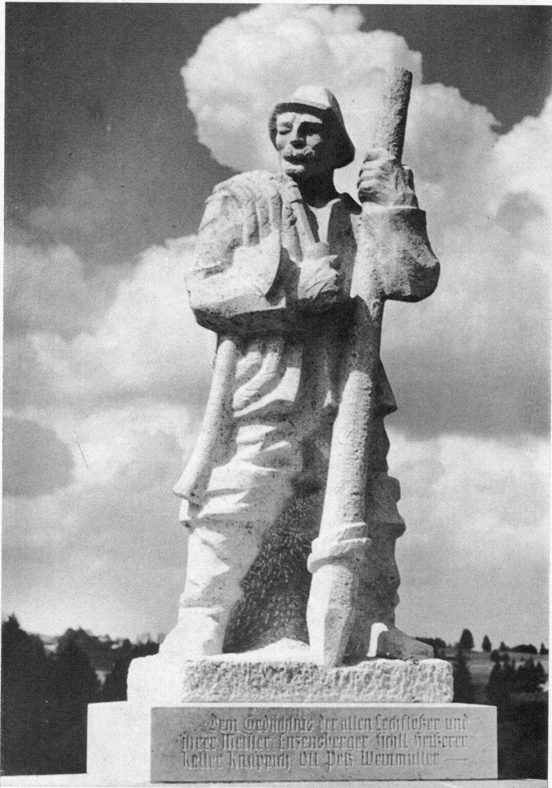 Eine steinerne Statue eines Mannes in schwerer Kleidung, der einen Stab in der Hand hält, steht auf einem Sockel mit einer Inschrift, die vielen Denkmale oder Marterln ähnelt, die in der Region zu finden sind; ein wolkenverhangener Himmel bildet den Hintergrund.