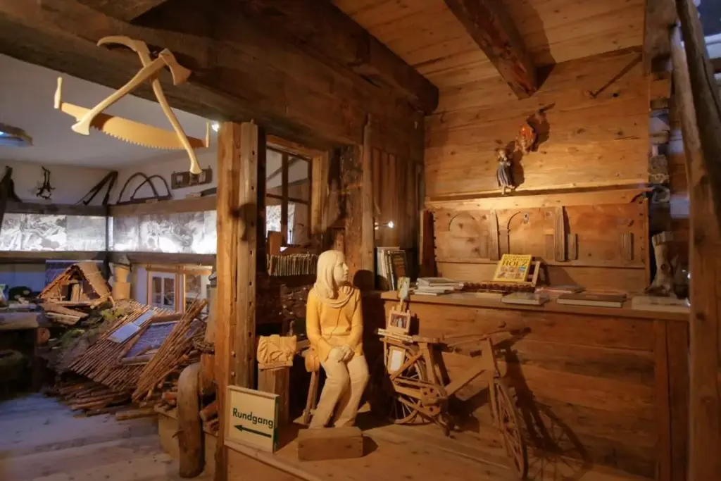 Das Tiroler Holzmuseum zeigt in einem rustikalen Holzinterieur traditionelle Werkzeuge, eine Schaufensterpuppe, die neben einem Spinnrad sitzt, und Informationstafeln in einer museumsähnlichen Ausstellung.