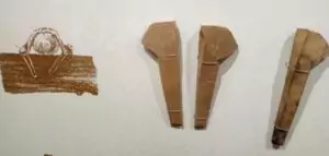 Drei uralte, braune, sargförmige Artefakte sind vertikal auf einer weißen Wand ausgestellt, begleitet von einem kleineren rechteckigen Objekt, bei dem es sich vermutlich um ein Flößerwerkzeug handelt und das verblasste Markierungen aufweist.