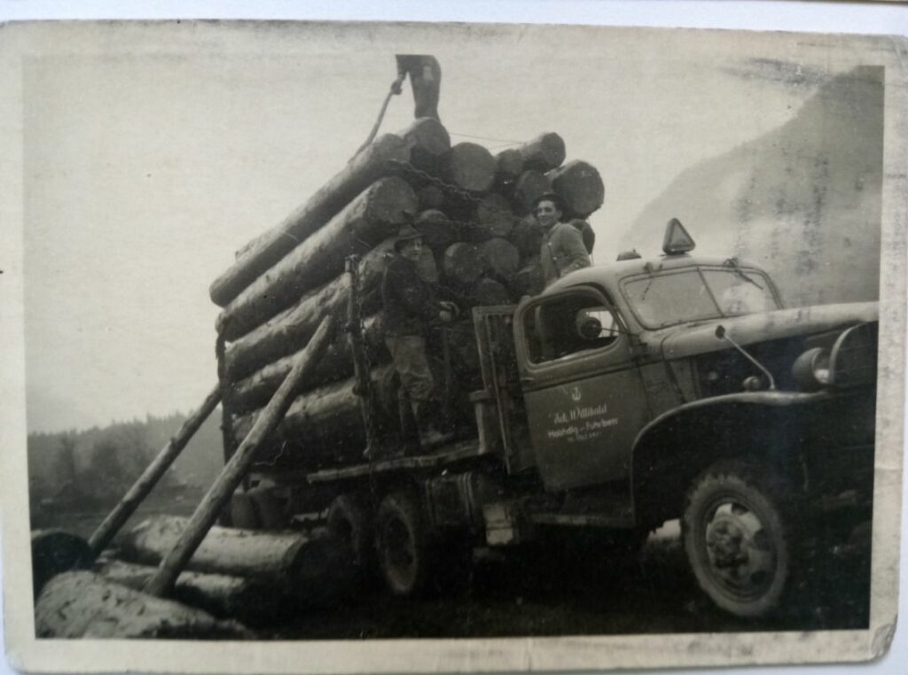 Schwarz-Weiß-Foto eines mit großen Baumstämmen beladenen Lastwagens; zwei Männer stehen neben und auf der Ladung, einer hält ein Werkzeug oder eine Stange.