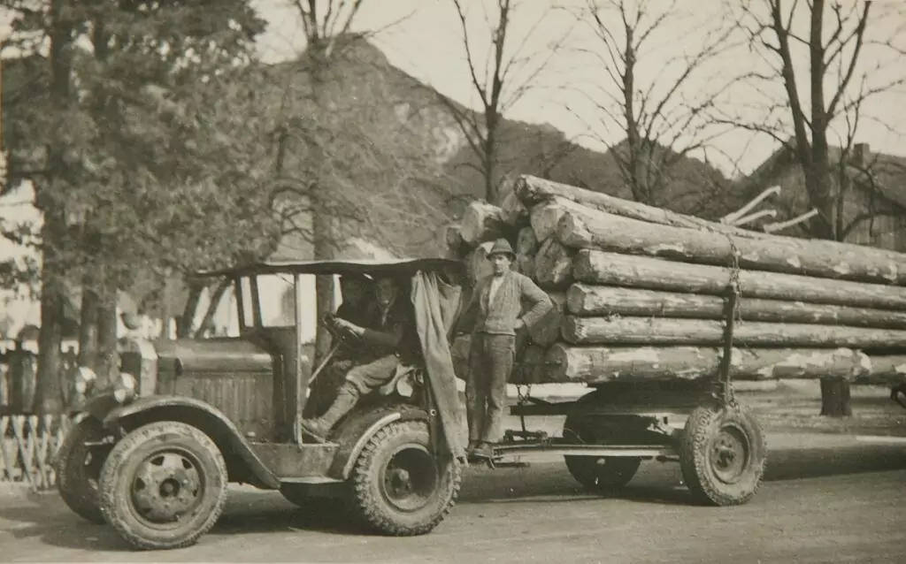 Ein Oldtimer-LKW mit einem Anhänger, der mit großen Baumstämmen beladen ist, zwei Männer stehen daneben, geparkt auf einer Straße mit Bäumen und einem Berg im Hintergrund.