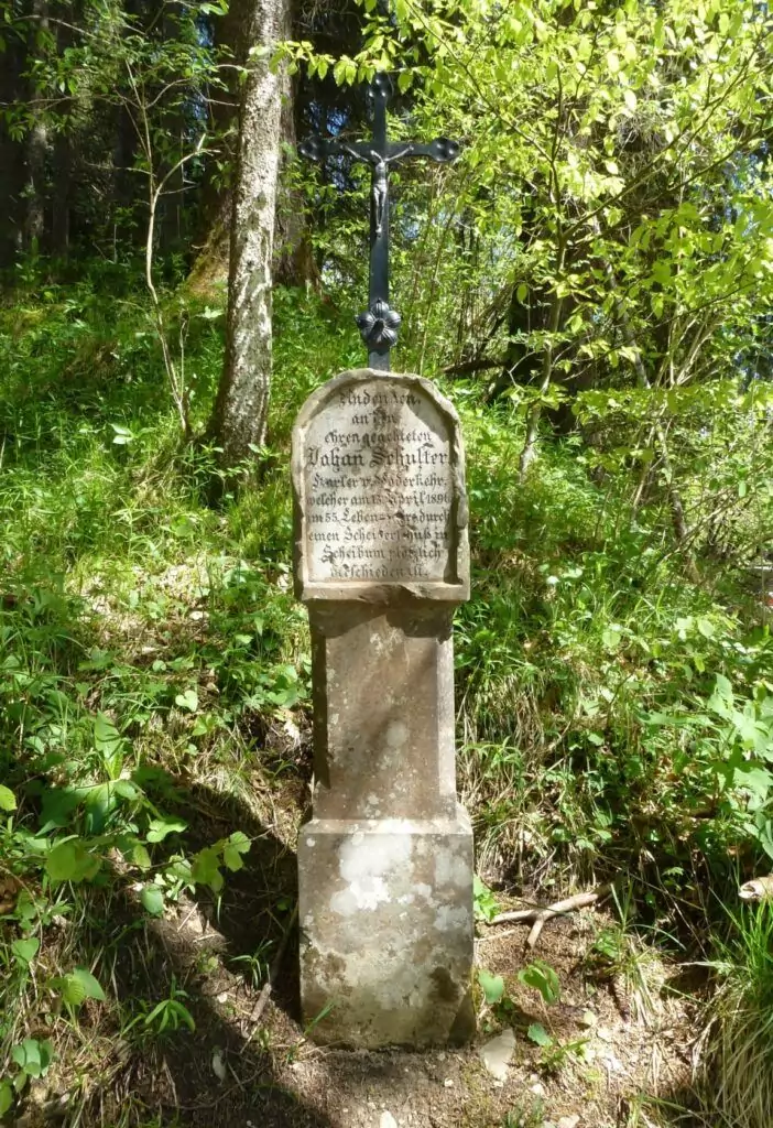 Das steinerne Denkmal mit dem Metallkreuz und der deutschen Inschrift ist ein Denkmale und steht auf einer Waldlichtung, umgeben von grünem Laub und Sonnenlicht.