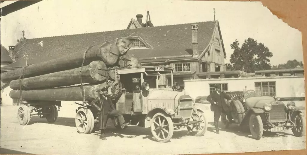 Ein mit großen Baumstämmen beladener Oldtimer-LKW ist neben einem Auto und mehreren Männern vor einem Gebäude mit Giebeldach geparkt.