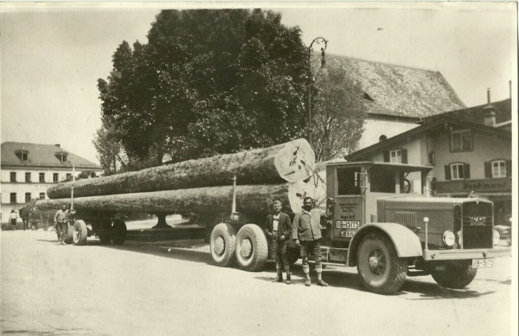 Schwarz-Weiß-Foto eines großen Lastwagens, der überlange Baumstämme transportiert, mit zwei daneben stehenden Männern in einer Stadtkulisse.
