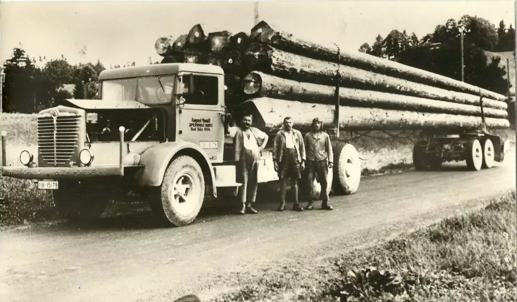Drei Männer stehen vor einem alten, mit großen Baumstämmen beladenen Holztransporter auf einer Landstraße, mit Bäumen und Feldern im Hintergrund.