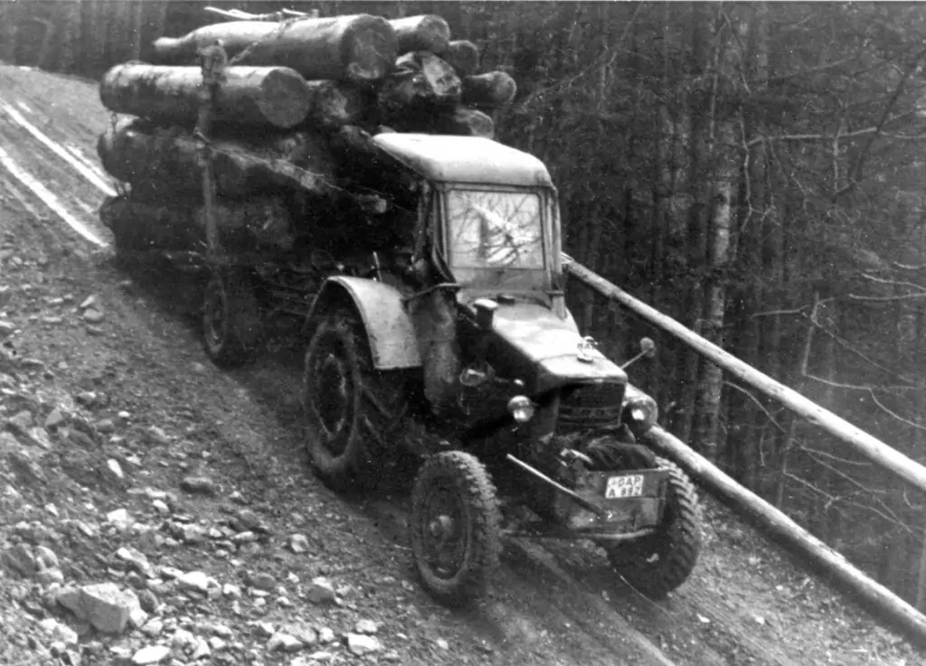 Ein mit großen Baumstämmen beladener Traktor fährt einen steilen, nicht asphaltierten Waldweg neben einer hölzernen Leitplanke hinunter.