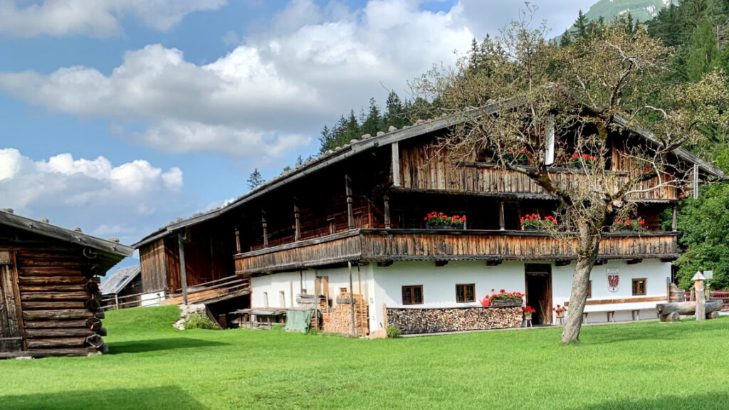 Ein traditionelles Alpenhaus aus Holz mit Blumenkästen, einem Baum davor und einem gepflegten grünen Rasen - das Heimatmuseum Sixenhof liegt vor bewaldeten Hügeln und einem teilweise bewölkten Himmel.