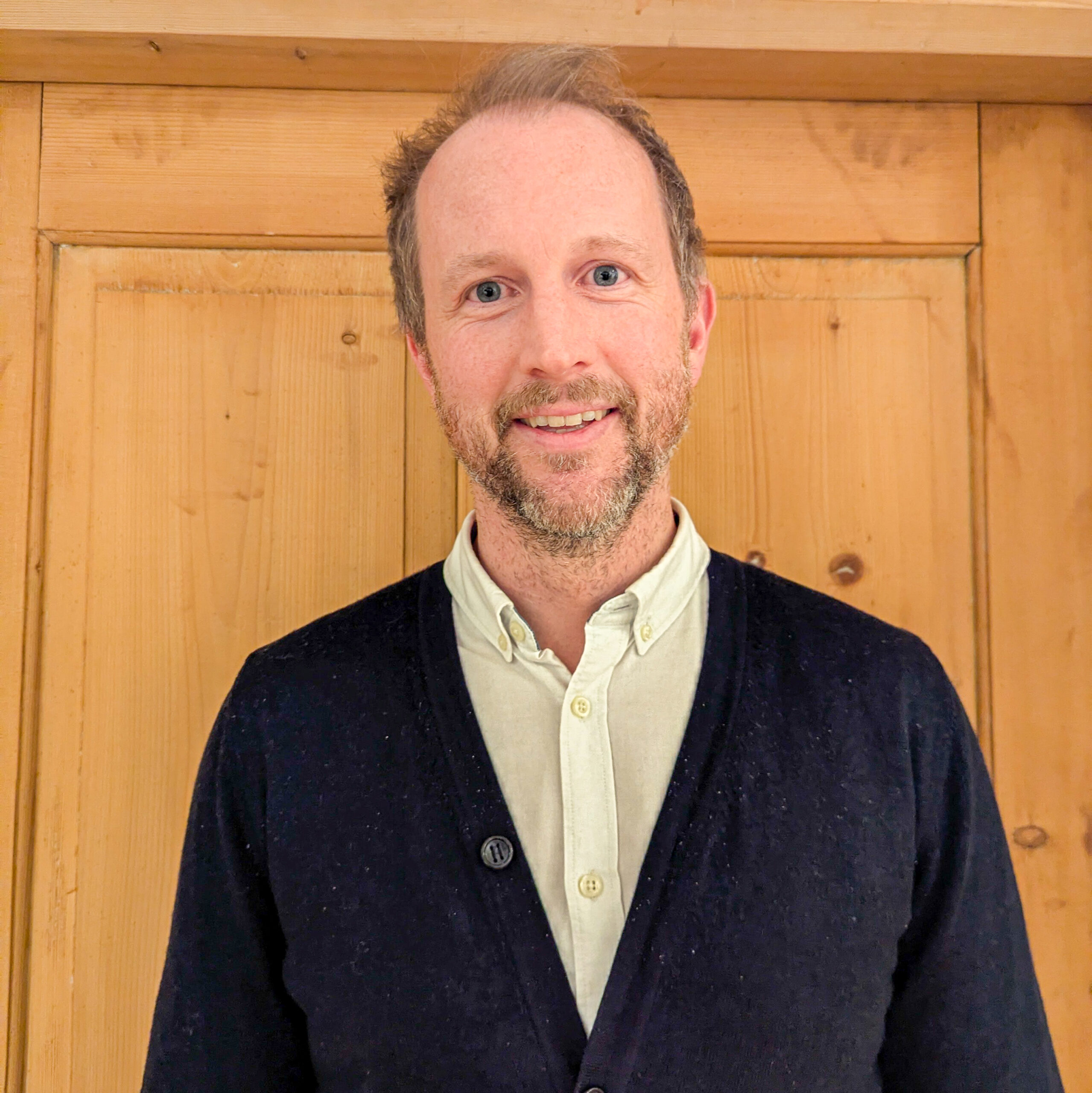 Felix Lassacher, ein Mann mit kurzen hellbraunen Haaren und einem Bart, der ein helles Hemd und eine dunkle Strickjacke trägt, steht vor einer Holztür und lächelt in die Kamera.