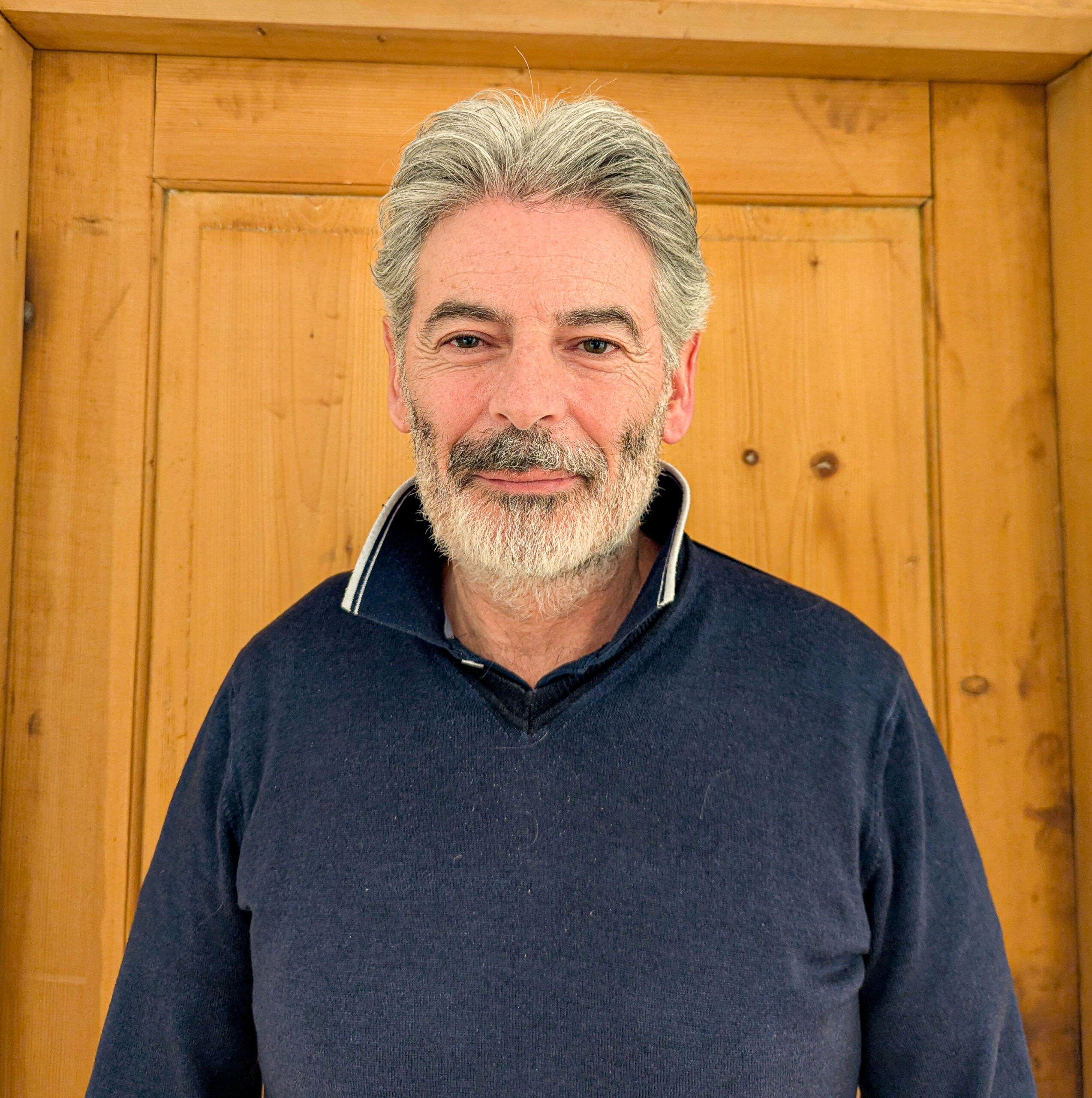 Ein Mann mittleren Alters mit grauen Haaren und Bart, der einen dunkelblauen Kragenpullover trägt, steht vor einer Holztür - es ist Christoph Stock.