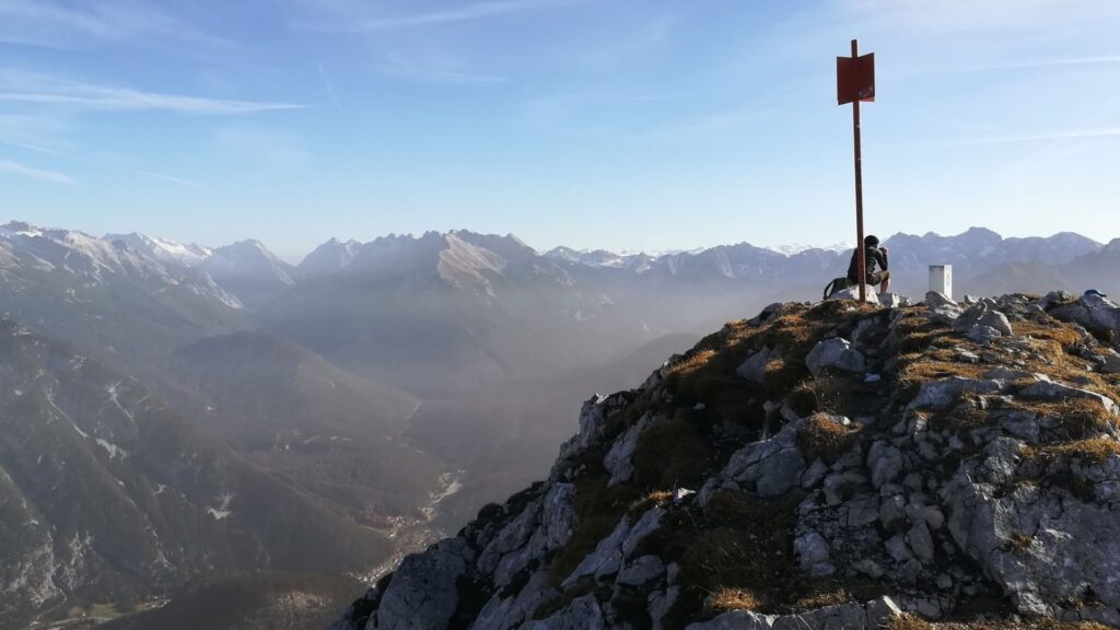 Eine Person mit einem Fahrrad sitzt auf einem felsigen Berggipfel in der Nähe eines Wegweisers und überblickt die fernen, dunstigen Berge der Arnspitze unter einem klaren Himmel.