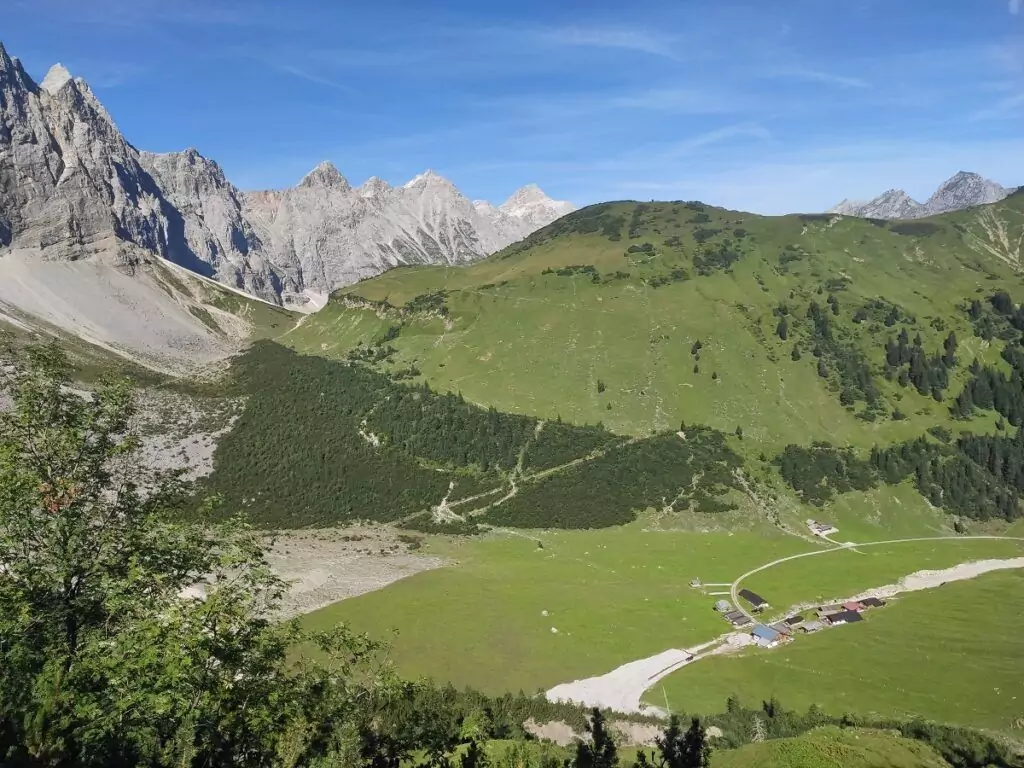 Eine Gebirgslandschaft mit felsigen Gipfeln im Hintergrund, grünen Hügeln und Wiesen, einer kurvenreichen Straße und einer kleinen Ansammlung von Gebäuden im Tal - der perfekte Lebensraum für den Bergpieper, der zwischen den Hügeln gesichtet werden kann.